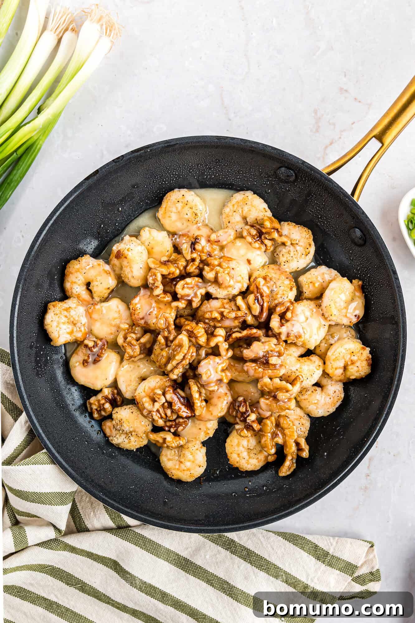 A skillet showcasing a harmonious blend of sautéed shrimp and walnuts, beautifully complemented by a scattering of fresh green onions and a rustic striped cloth laid beside it.