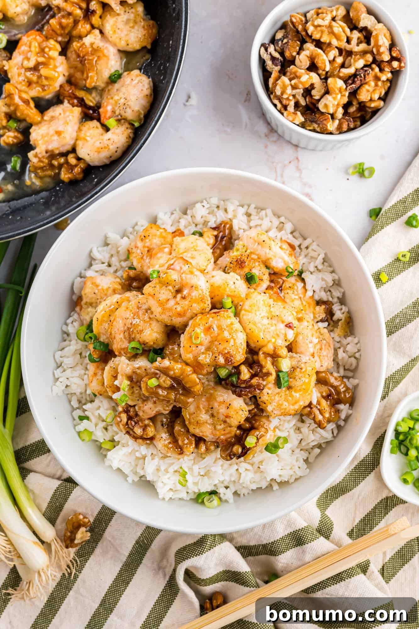A beautifully presented bowl of rice, generously topped with an array of crispy shrimp and crunchy walnuts. The dish is elegantly garnished with finely chopped green onions, while a striped cloth, a separate bowl of walnuts, and more fresh green onions are artfully arranged nearby, completing the inviting scene.