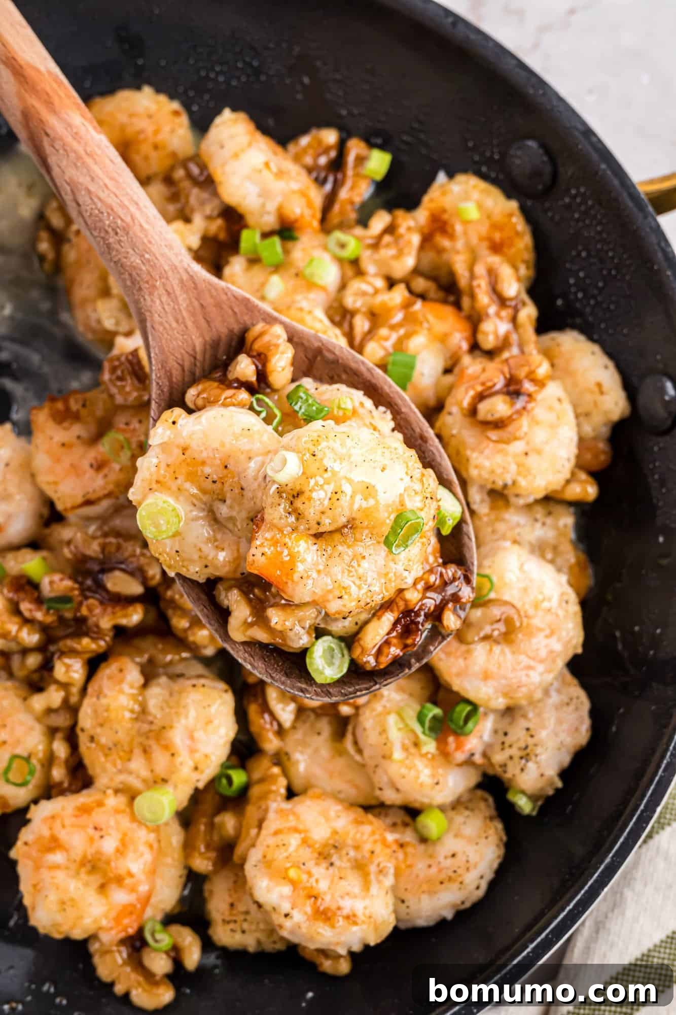 A wooden spoon delicately scoops a portion of Honey Walnut Shrimp, garnished with fresh green onions, directly from a sizzling skillet, invitingly ready for serving.