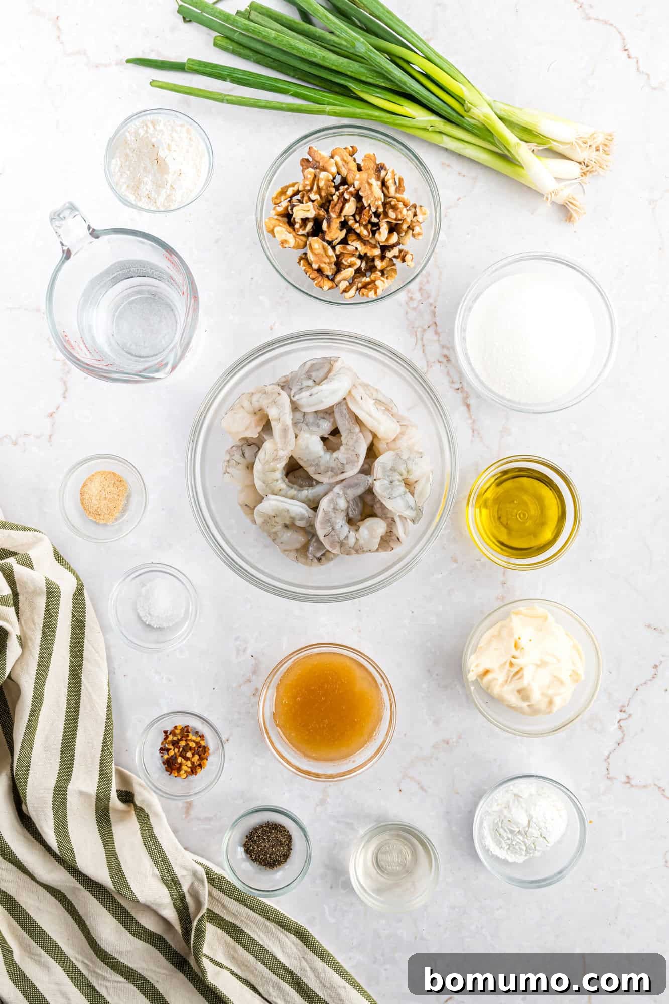 Various ingredients laid out on a kitchen countertop, including raw shrimp, whole walnuts, fresh green onions, and an assortment of liquids and powders in small bowls, all resting beside a neatly folded striped cloth.