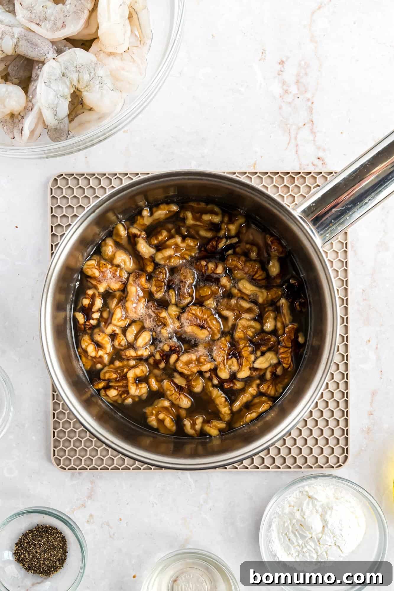 A close-up view of walnuts gently simmering in a clear, sugary liquid inside a small saucepan. The saucepan is situated on a kitchen countertop, surrounded by other raw ingredients like fresh shrimp and various seasonings, creating a dynamic culinary scene.