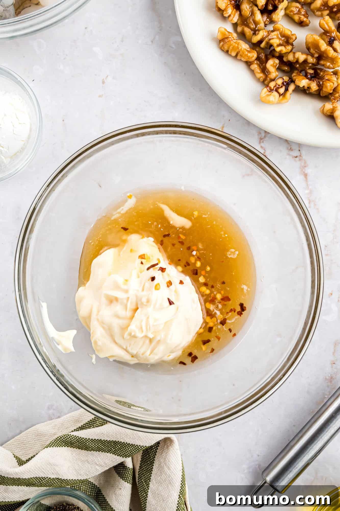 A clear glass bowl holding a blend of mayonnaise, golden honey, and vibrant red pepper flakes, poised on a polished marble surface. Nearby, a plate of candied walnuts and a striped kitchen cloth complete the inviting tableau.