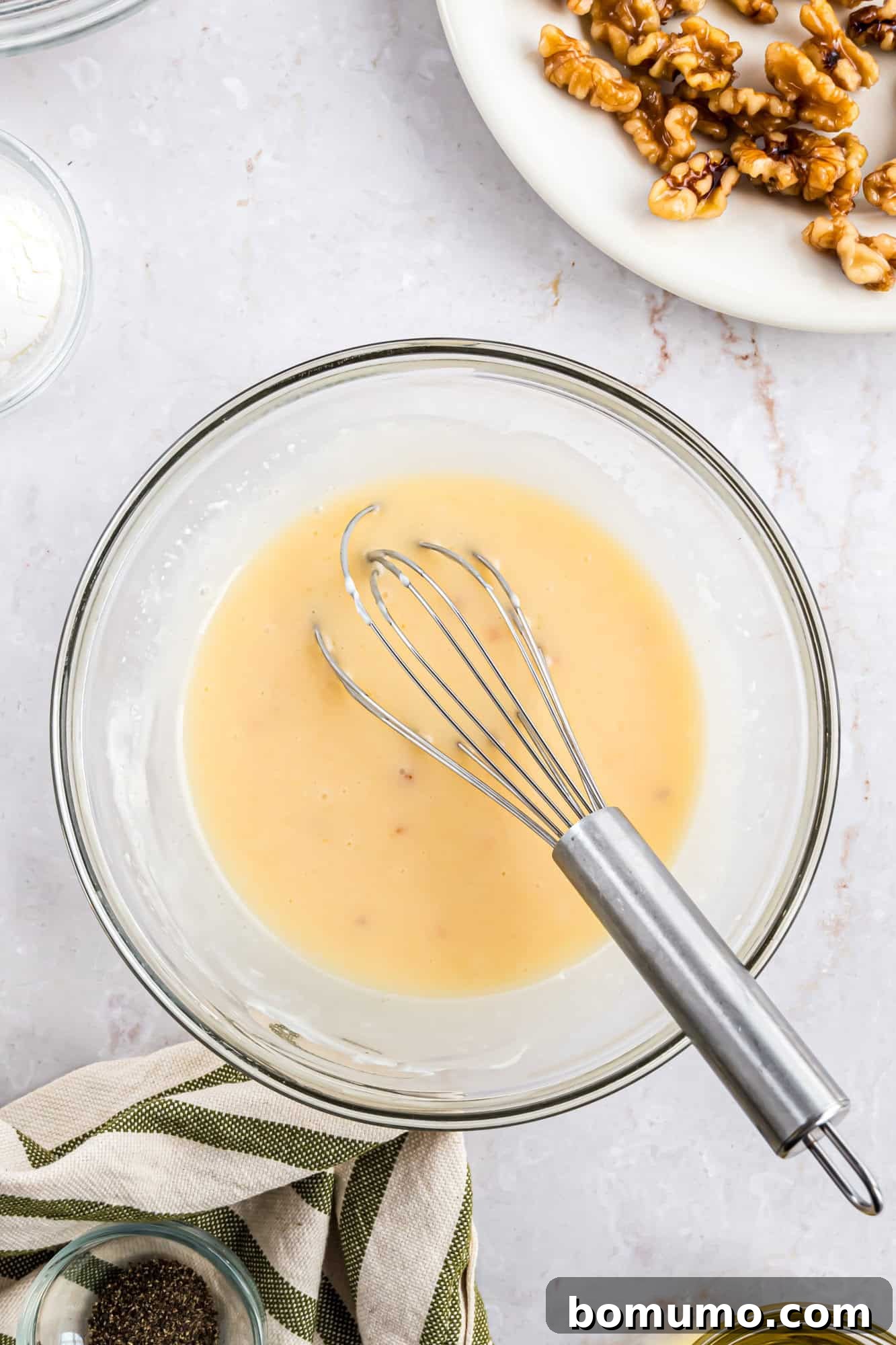 A whisk gracefully blending a creamy mixture within a glass bowl, positioned on a gleaming marble surface. Adjacent to it, a plate of freshly prepared walnuts and a striped kitchen cloth hint at the culinary process underway.