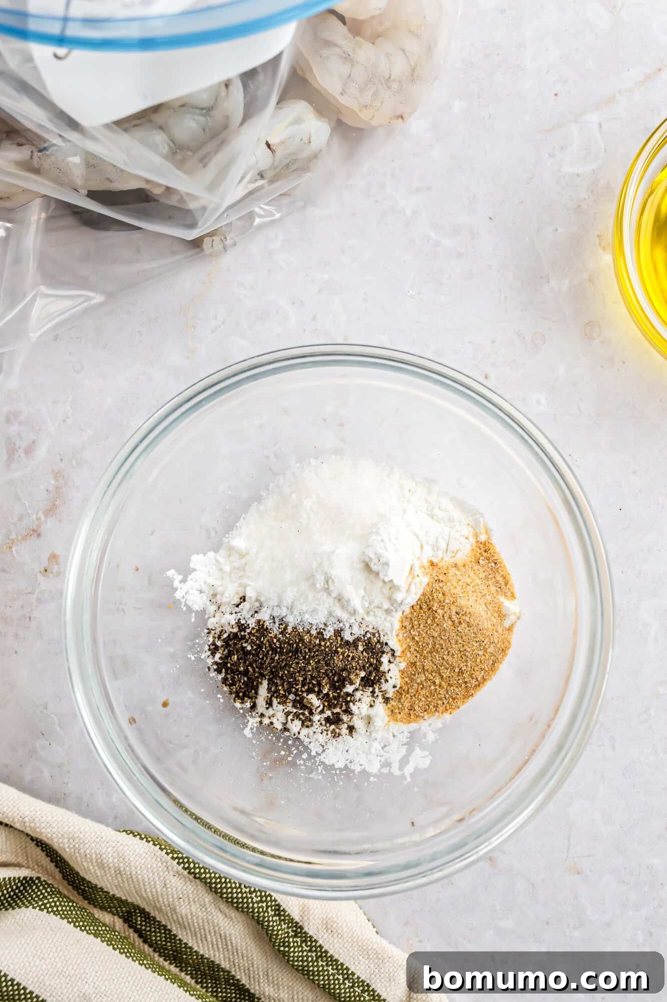 A glass bowl containing a mix of salt, pepper, and garlic powder rests on a pristine marble surface. A partial view shows avocado oil in a small container, raw shrimp ready for preparation in a plastic bag, and a striped cloth nearby, all set for cooking.