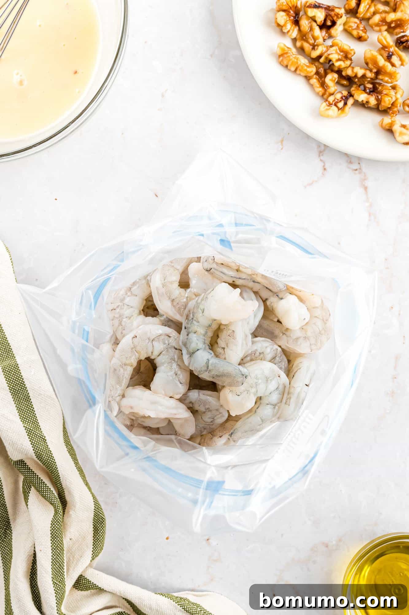 A gallon-size resealable bag filled with raw, uncooked shrimp is placed on a sleek marble surface, flanked by a striped kitchen cloth, a bowl of liquid ingredients, a plate of walnuts, and a small bottle of cooking oil, signifying the start of a meal preparation.