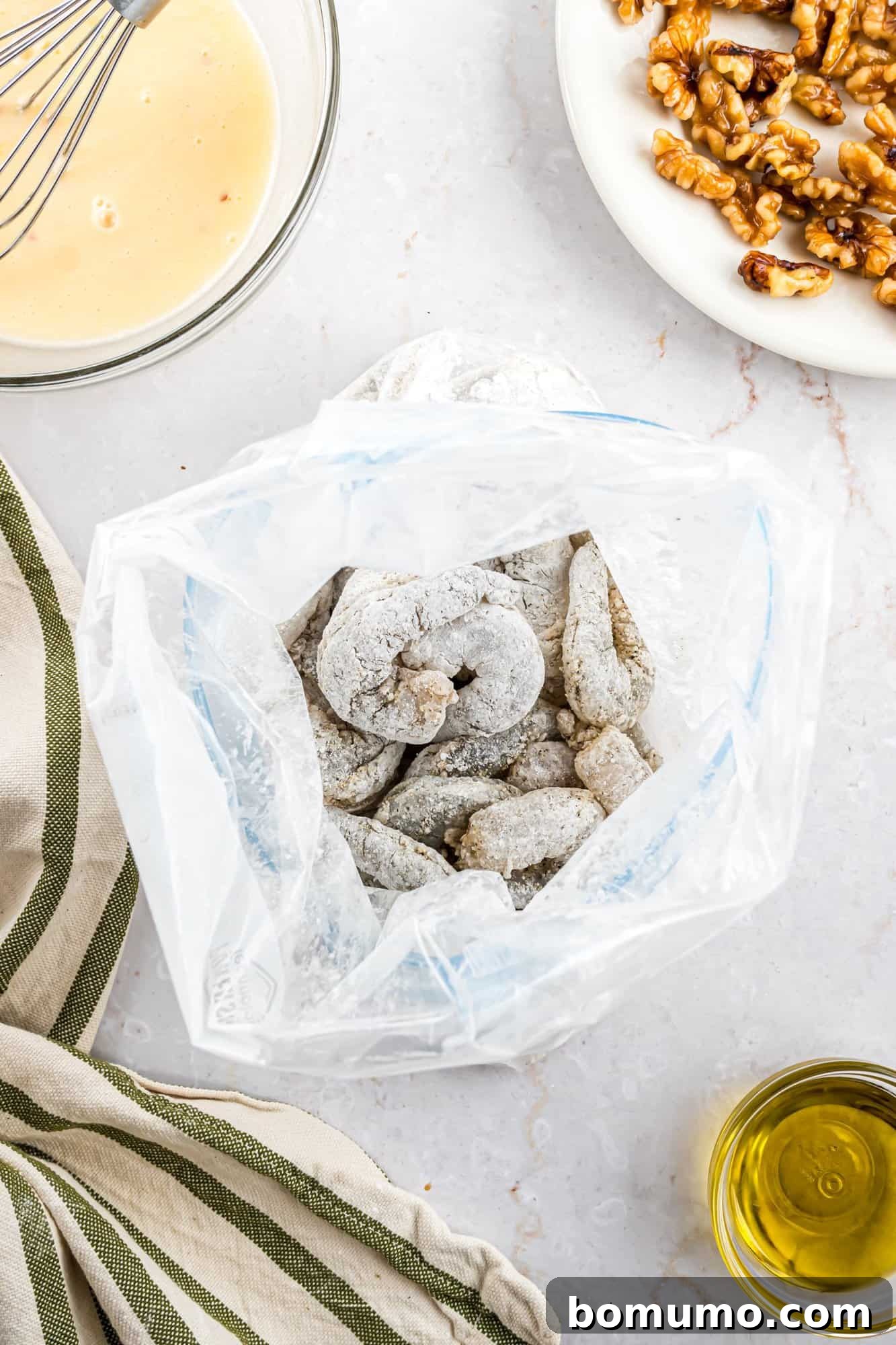Raw shrimp, perfectly coated in a light dusting of flour, rests inside a plastic bag on a polished marble surface. Beside it, a bowl filled with a creamy batter, a plate of walnuts, a striped kitchen cloth, and a small dish of olive oil are neatly arranged, indicating a delicious recipe in progress.