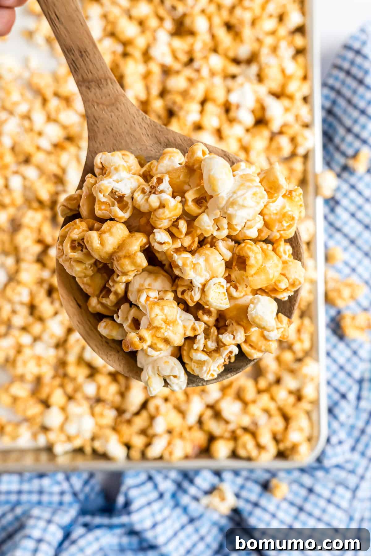A wooden spoon scooping freshly baked caramel corn from a sheet pan, showcasing its golden, crispy texture.