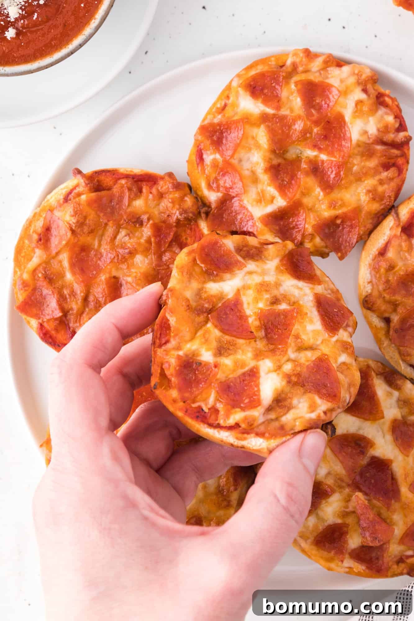 Hand holding a homemade bagel bite pizza with melted cheese and pepperoni.