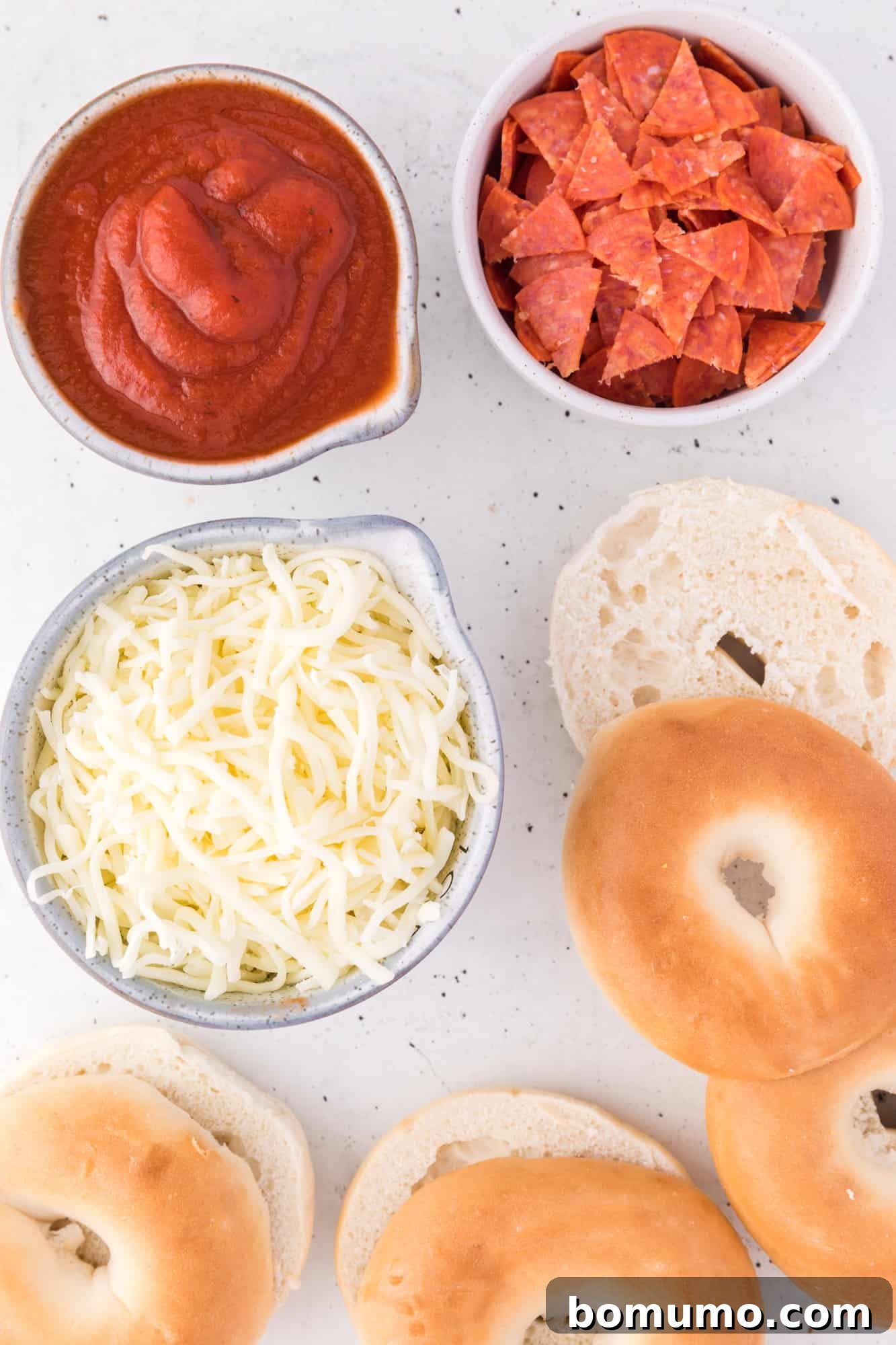 Overhead view of ingredients needed for bagel bite pizzas, including mini bagels, pizza sauce, mozzarella, and chopped pepperoni.