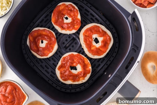 Mini bagel halves in air fryer, topped with a layer of savory pizza sauce.