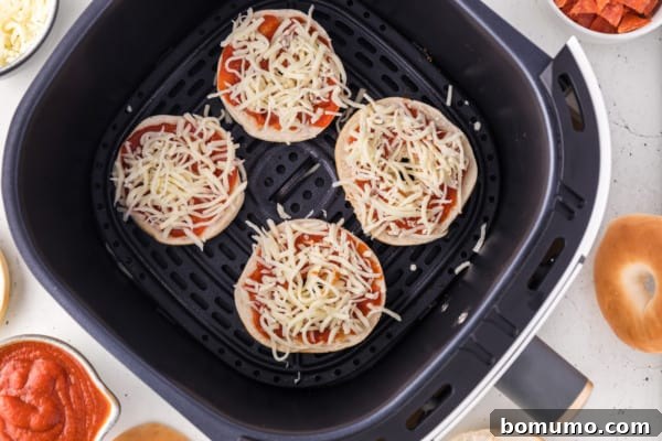 Mini bagel halves in air fryer, topped with pizza sauce and a generous layer of shredded mozzarella.