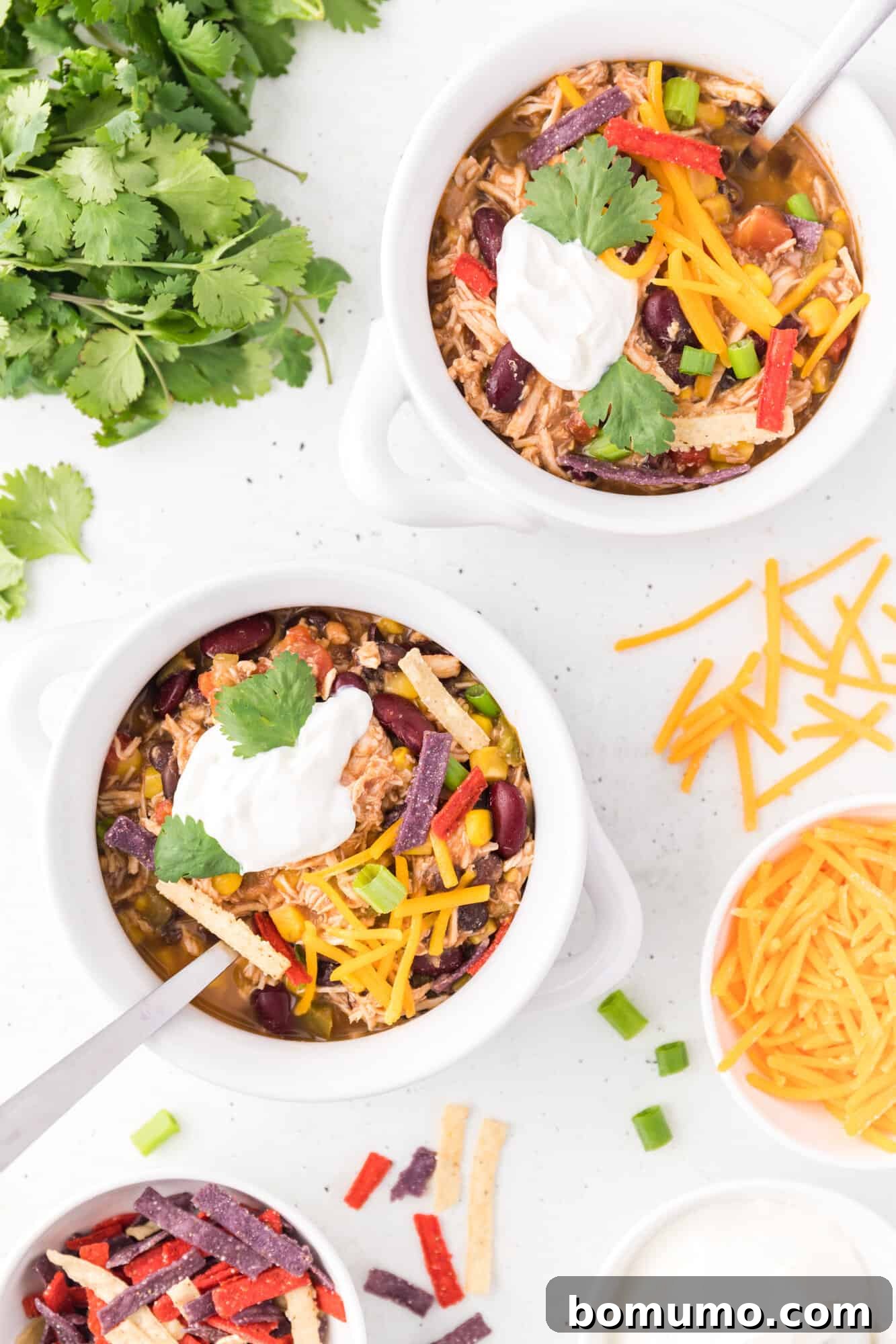 Overhead view of two bowls of chicken taco soup, garnished with sour cream and tortilla strips, with fresh cilantro and bowls of additional toppings arranged on a kitchen countertop.