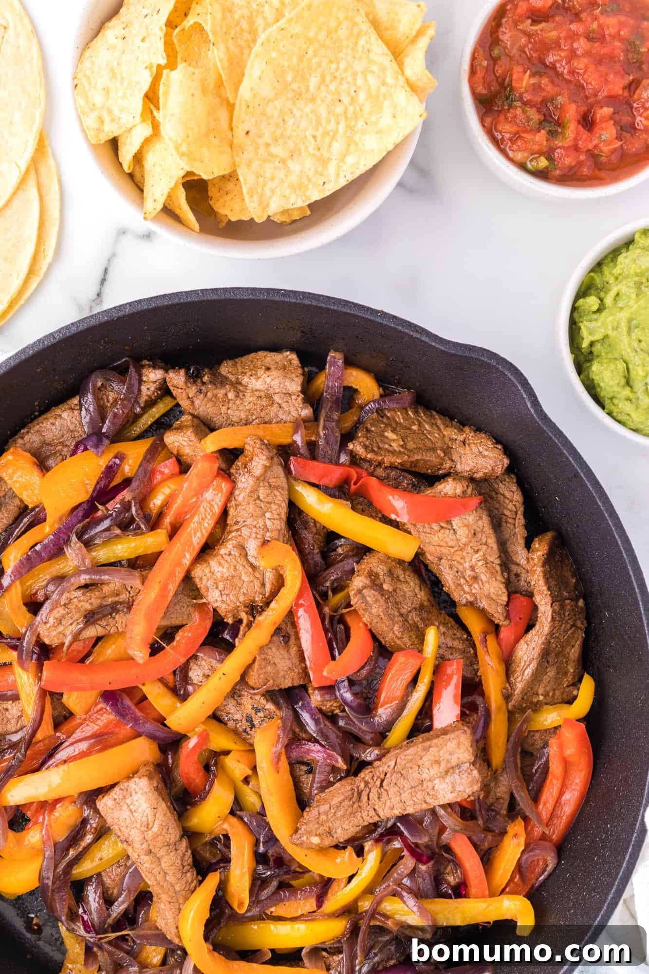Overhead view of sizzling steak fajitas in a cast-iron skillet, perfectly cooked, surrounded by bowls of fresh tortilla chips, tangy salsa, and creamy guacamole, all ready for a delicious Tex-Mex meal.