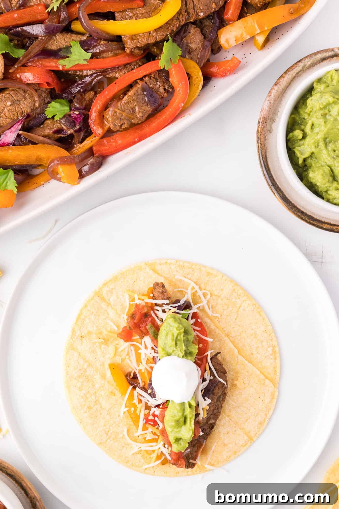 Overhead view of a perfectly assembled steak fajita on a plate, featuring tender steak, vibrant vegetables, and rich garnishes, artfully positioned next to a serving dish filled with more sizzling steak fajitas, creating an inviting presentation.