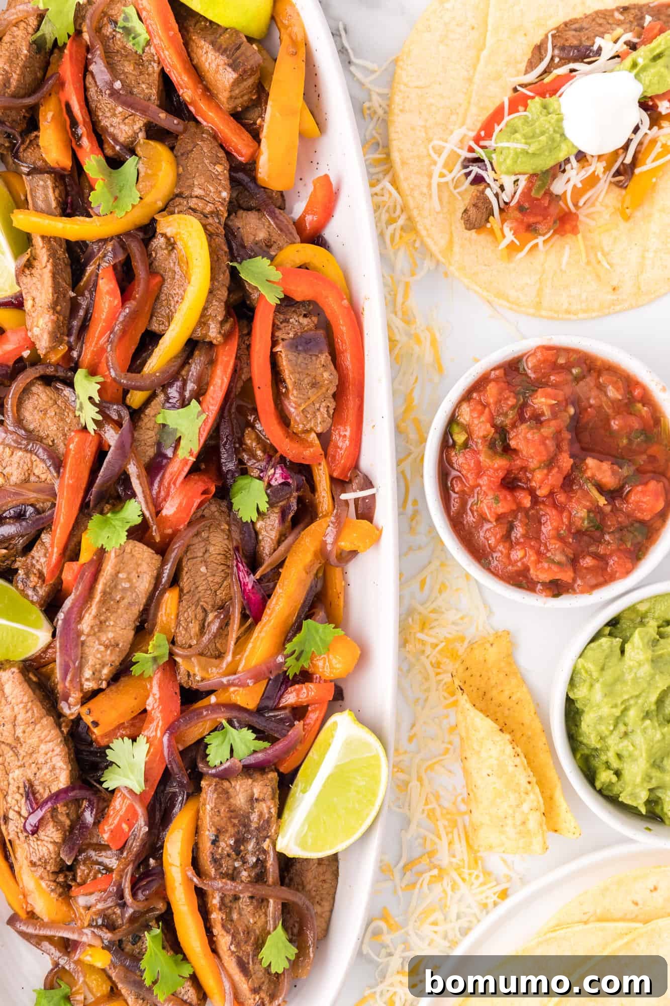 Overhead view of a long, elegant serving dish filled with perfectly cooked steak fajitas, beautifully garnished with fresh lime wedges and vibrant chopped cilantro, presented alongside inviting bowls of various toppings and an already assembled fajita on a plate, creating a stunning and complete meal display.