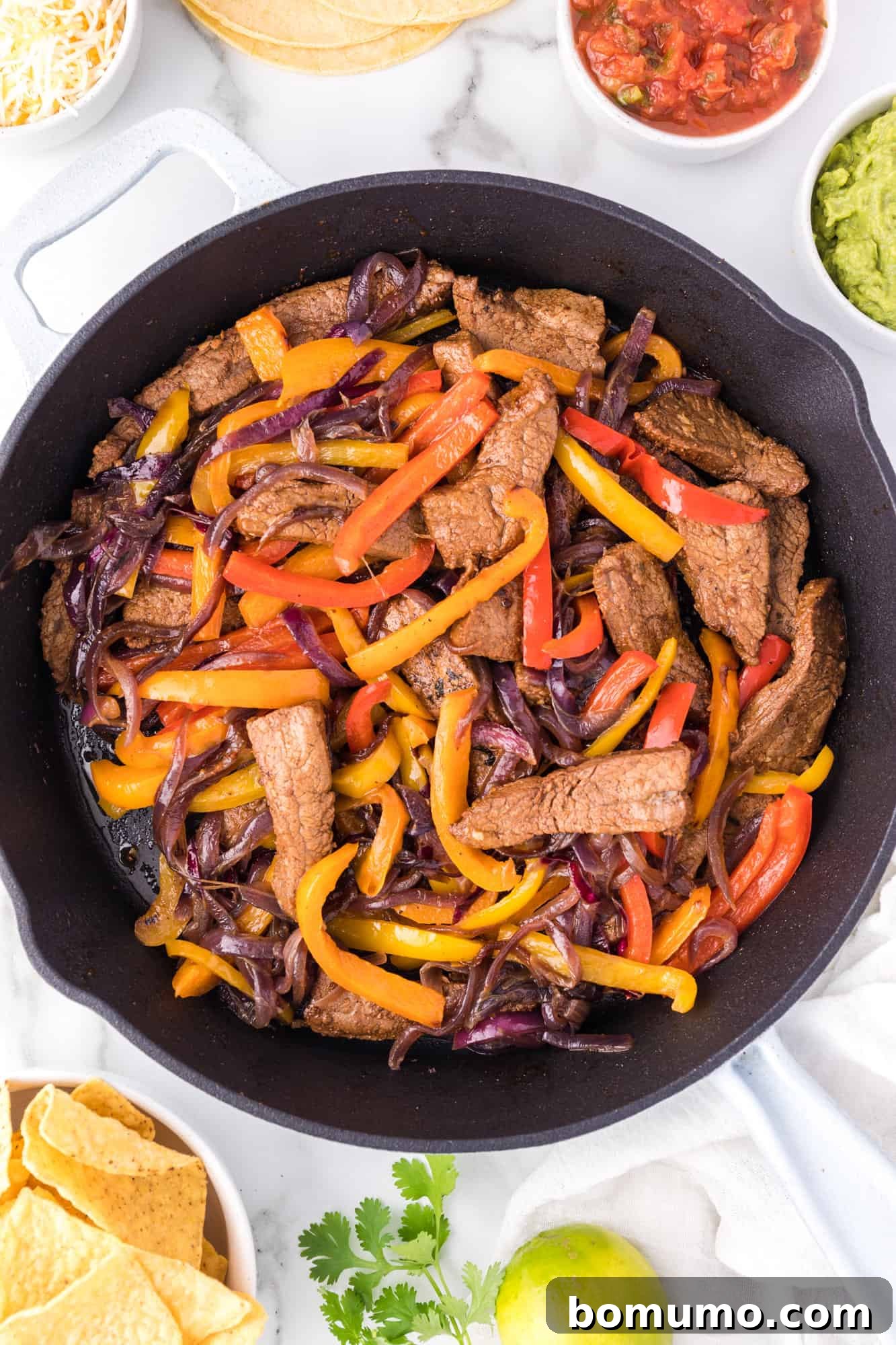 Overhead view of perfectly cooked steak fajitas in a large skillet, beautifully arranged and surrounded by an enticing spread of fresh toppings in small bowls, including guacamole, salsa, sour cream, and shredded cheese.