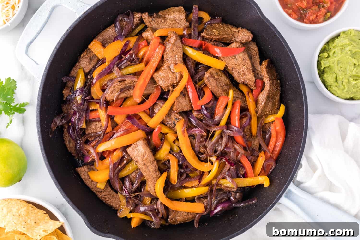 Overhead view of a bustling dinner setting featuring sizzling steak fajitas in a cast-iron skillet, positioned alongside inviting bowls of crunchy tortilla chips, fresh salsa, and creamy guacamole, ready for a convivial meal.