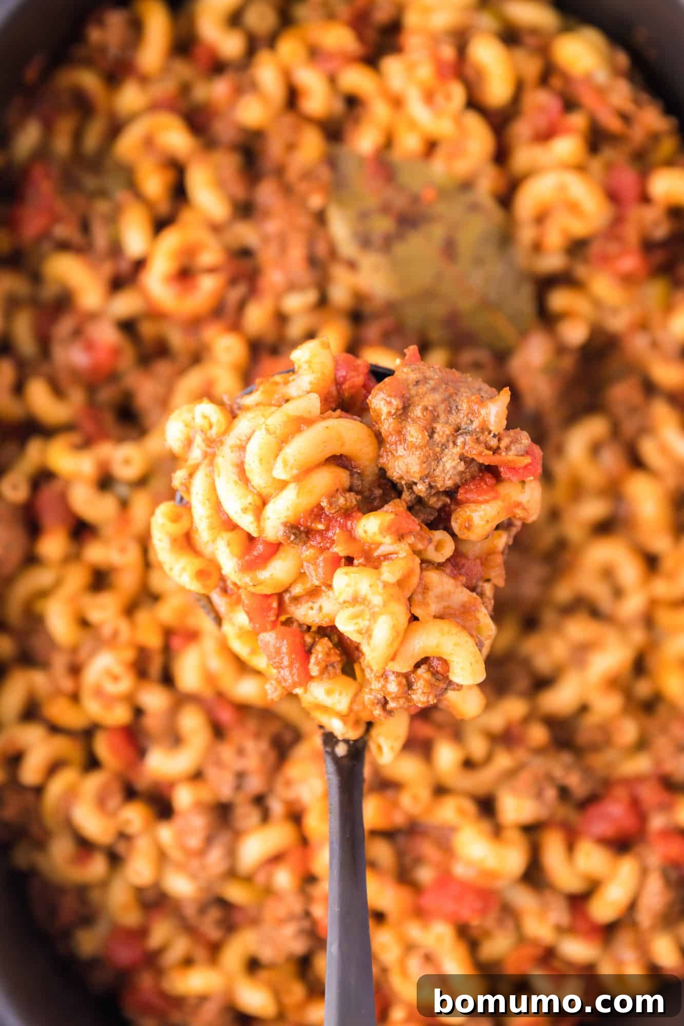 Overhead view of a large spoonful of American goulash held over the slow cooker, highlighting the rich tomato sauce and tender macaroni.