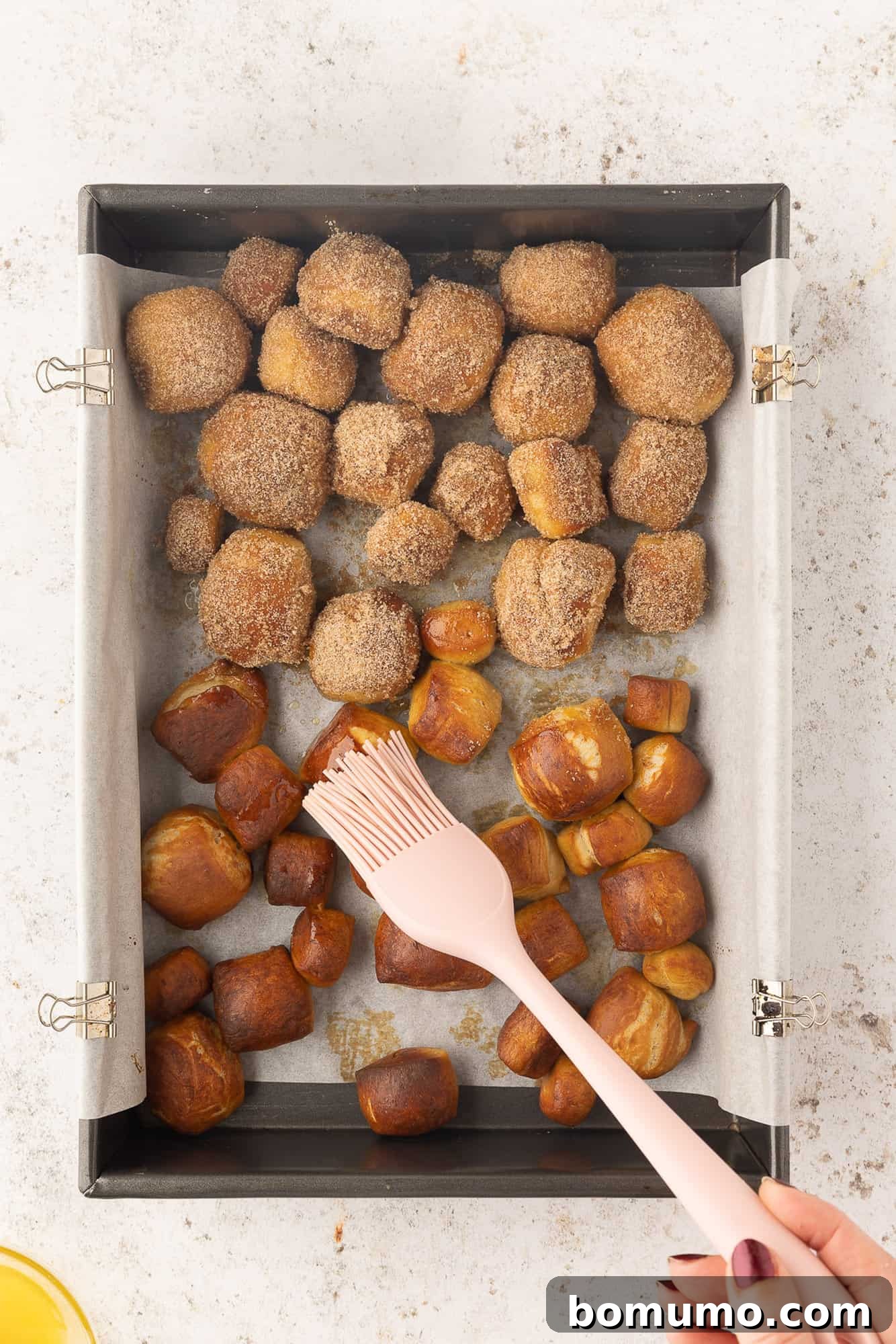 Baked pretzel bites on a baking sheet being brushed with butter, next to pretzel bites covered in cinnamon sugar.