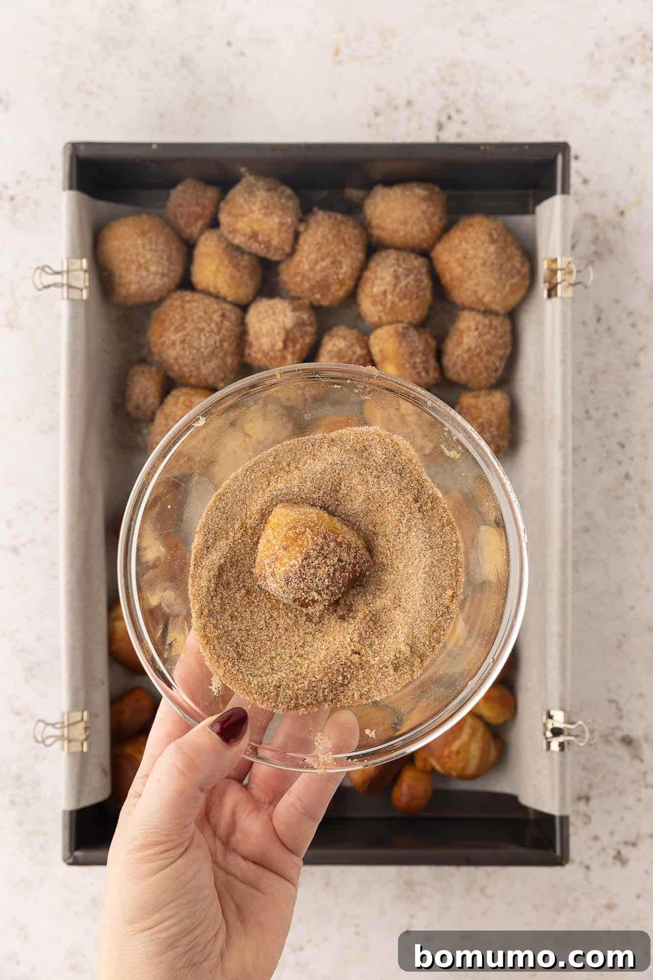 A hand holding a bowl of cinnamon sugar with a partially coated pretzel bite, over more pretzel bites on a baking sheet.