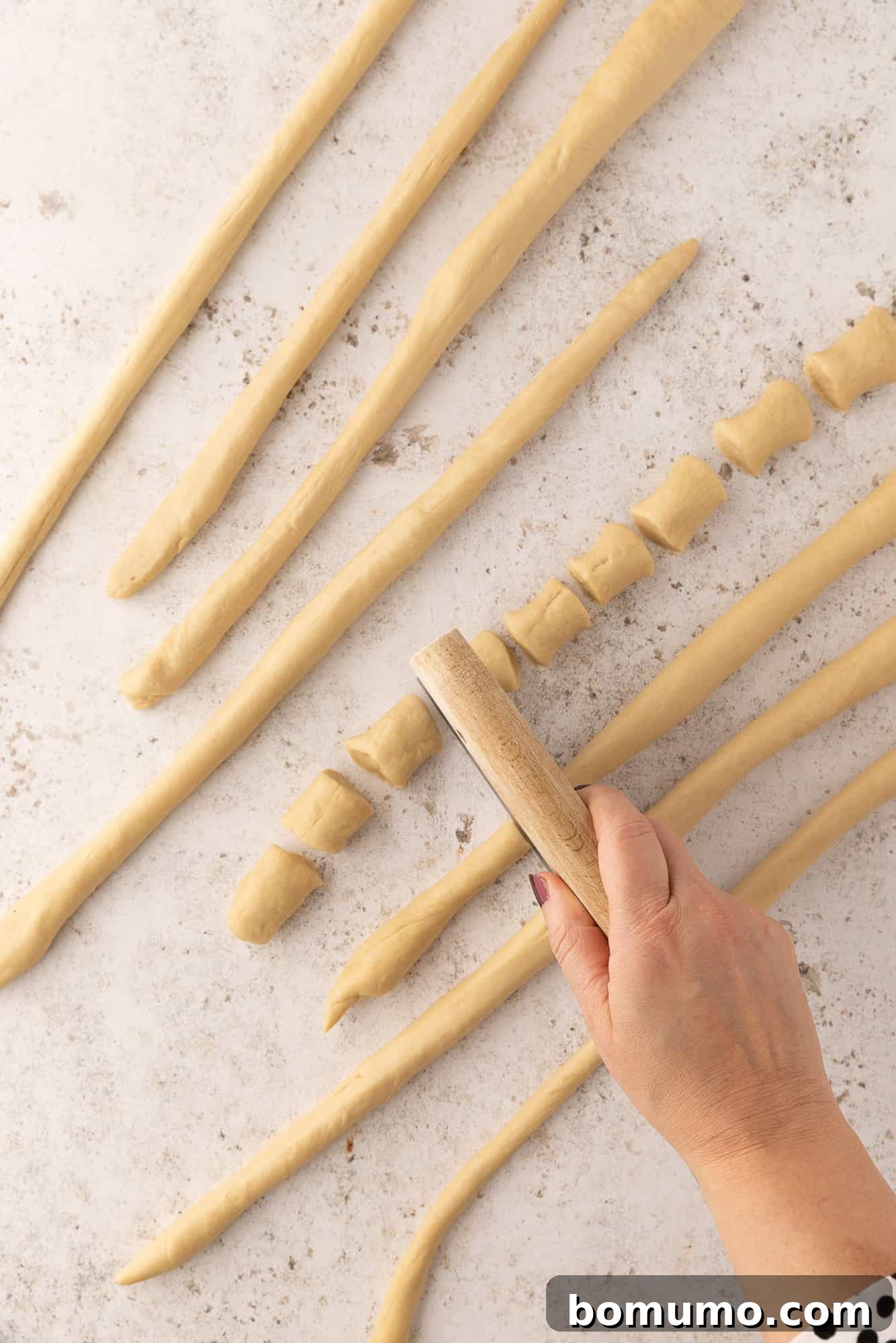A hand using a dough cutter to cut ropes of pretzel dough into small bite-sized pieces.