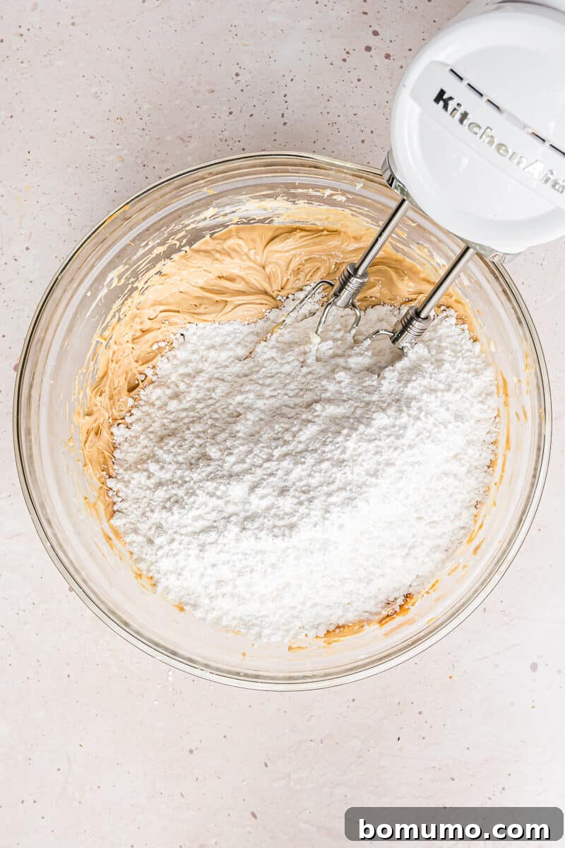 Powdered sugar being carefully and gradually added to the peanut butter and butter mixture in a mixing bowl.