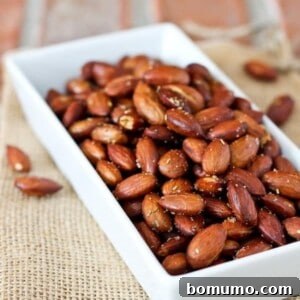 A rectangular white dish filled with rosemary roasted almonds sits on a piece of burlap, with a few almonds scattered around.