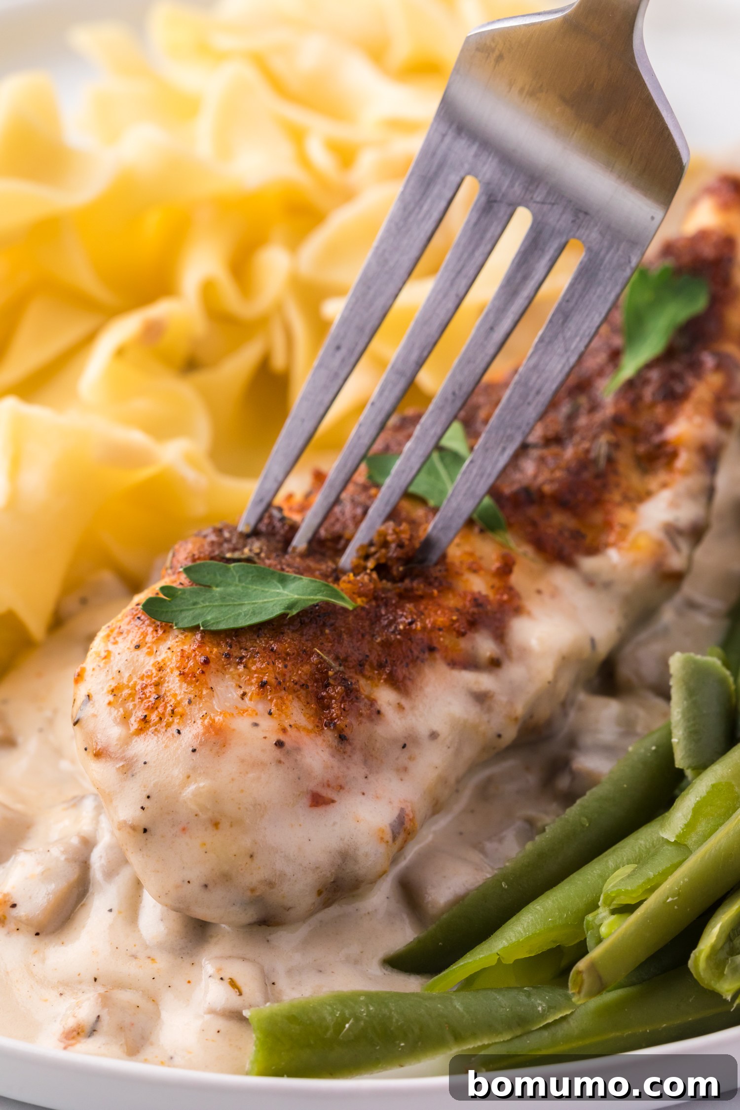 Close up of a fork stuck into a piece of cream of mushroom chicken served over egg noodles next to green beans on a white plate, ready to be enjoyed.