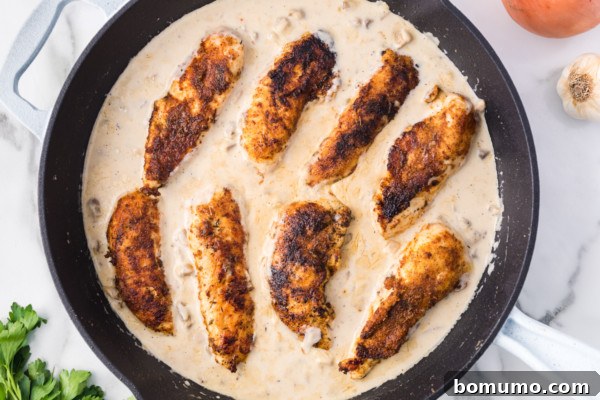 Overhead view of browned chicken tenders nestled back into a rich, creamy mushroom gravy in a skillet, garnished with fresh herbs.