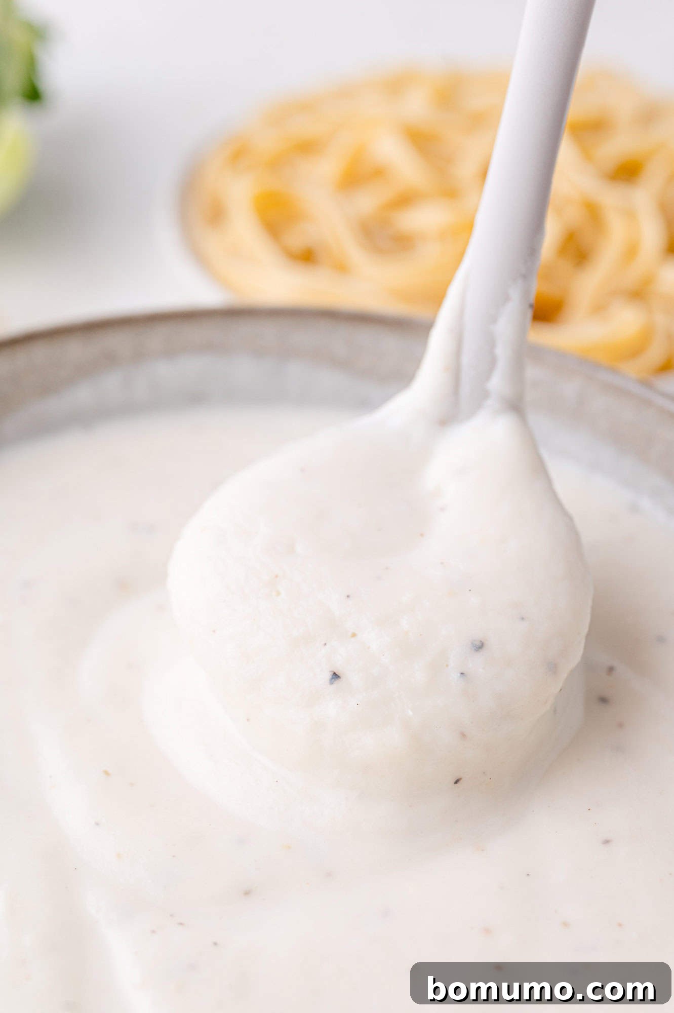 A ladle scoops creamy white Alfredo sauce from a bowl, with some fettuccine pasta visible in the background.