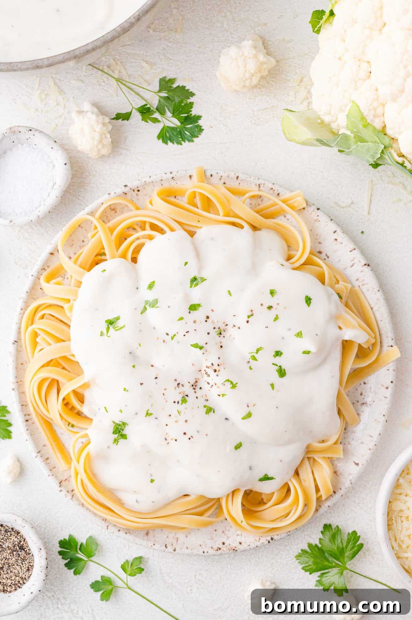 A plate of fettuccine pasta topped with creamy white sauce, garnished with black pepper and parsley. Surrounding the plate are small bowls and parsley leaves on a light surface.