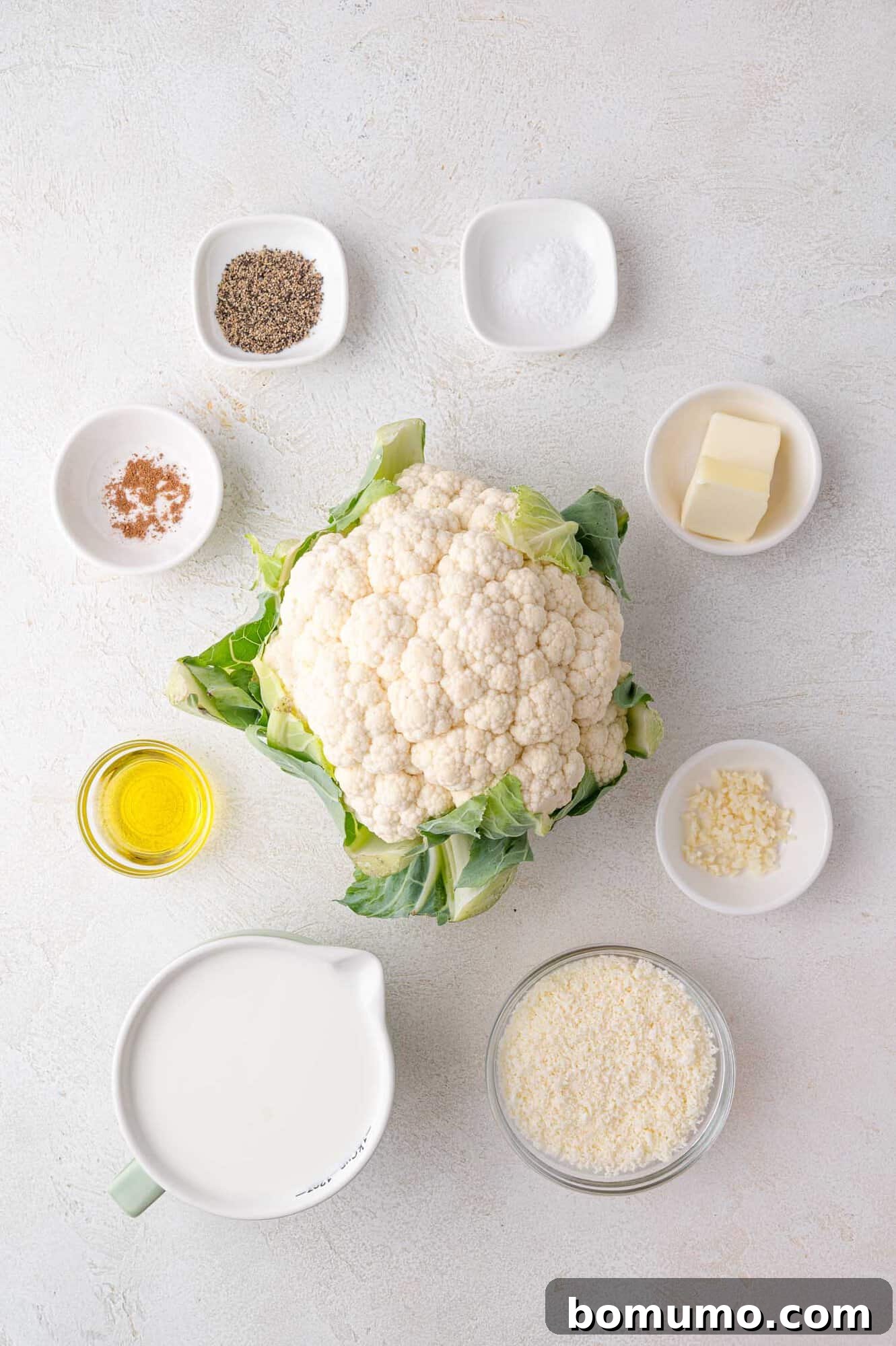 Top-down view of cauliflower surrounded by bowls containing milk, olive oil, butter, grated cheese, minced garlic, and spices on a white surface.