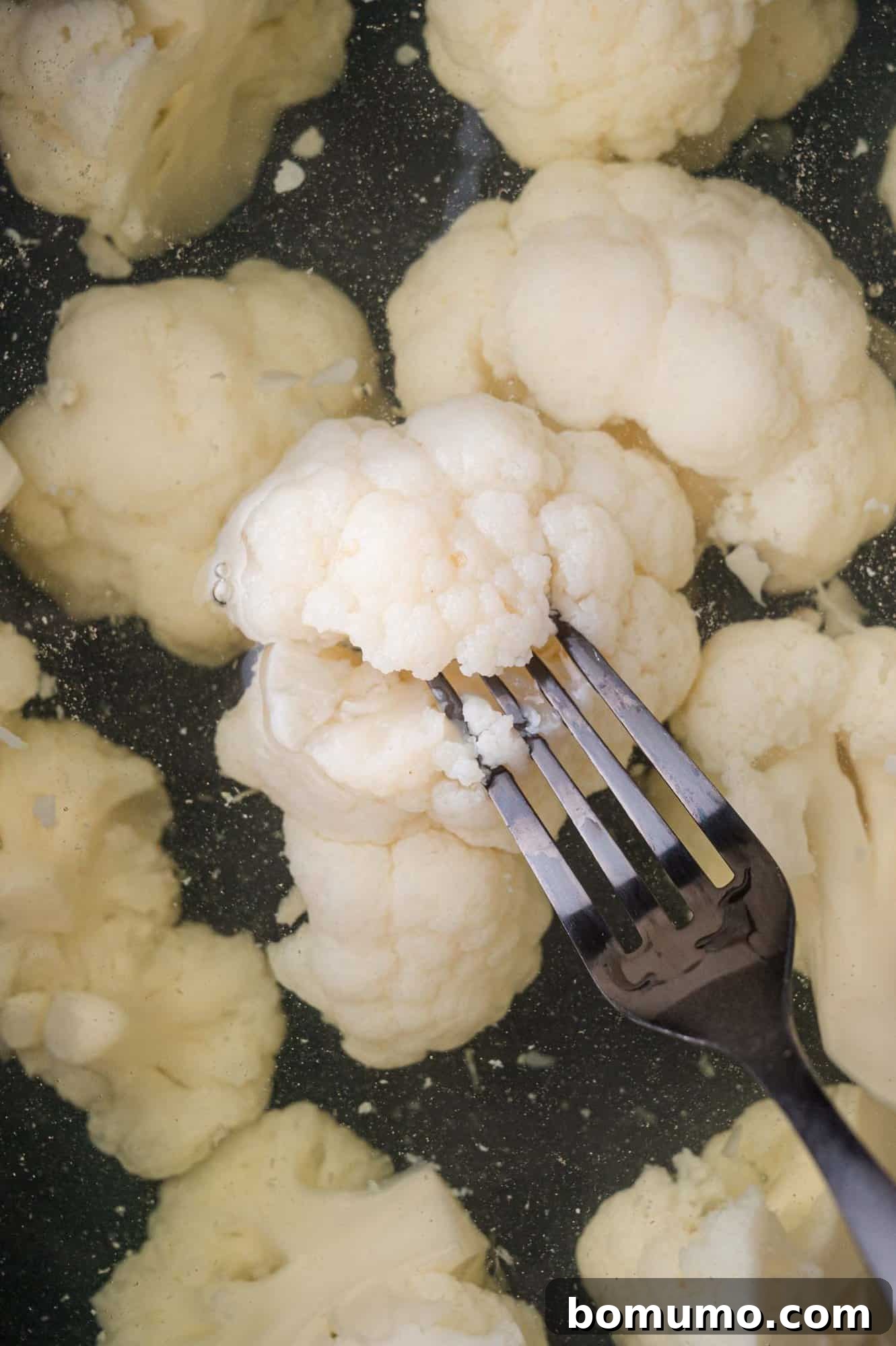 Close-up of a fork piercing a piece of cauliflower submerged in water, surrounded by other cauliflower pieces.