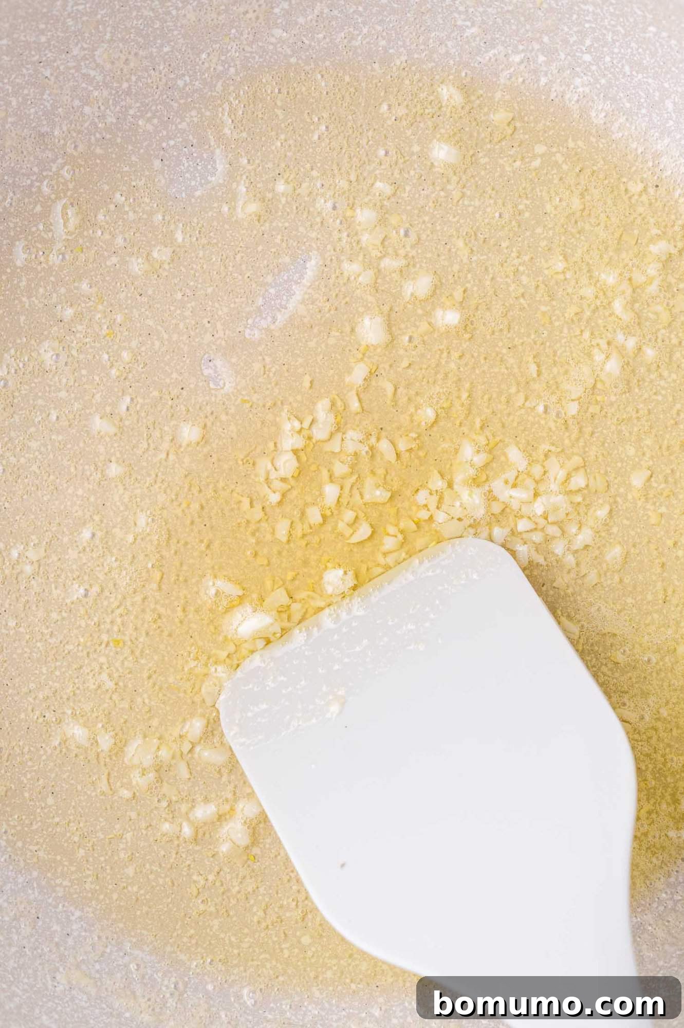 Chopped garlic sizzling in melted butter in a pan, stirred with a white spatula.