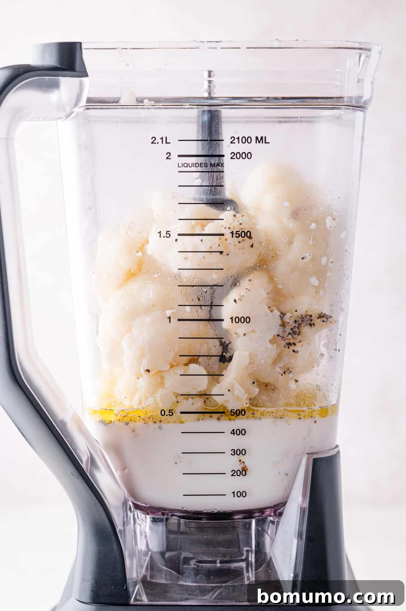 A blender filled with cauliflower, milk, herbs, and black pepper on a white surface.