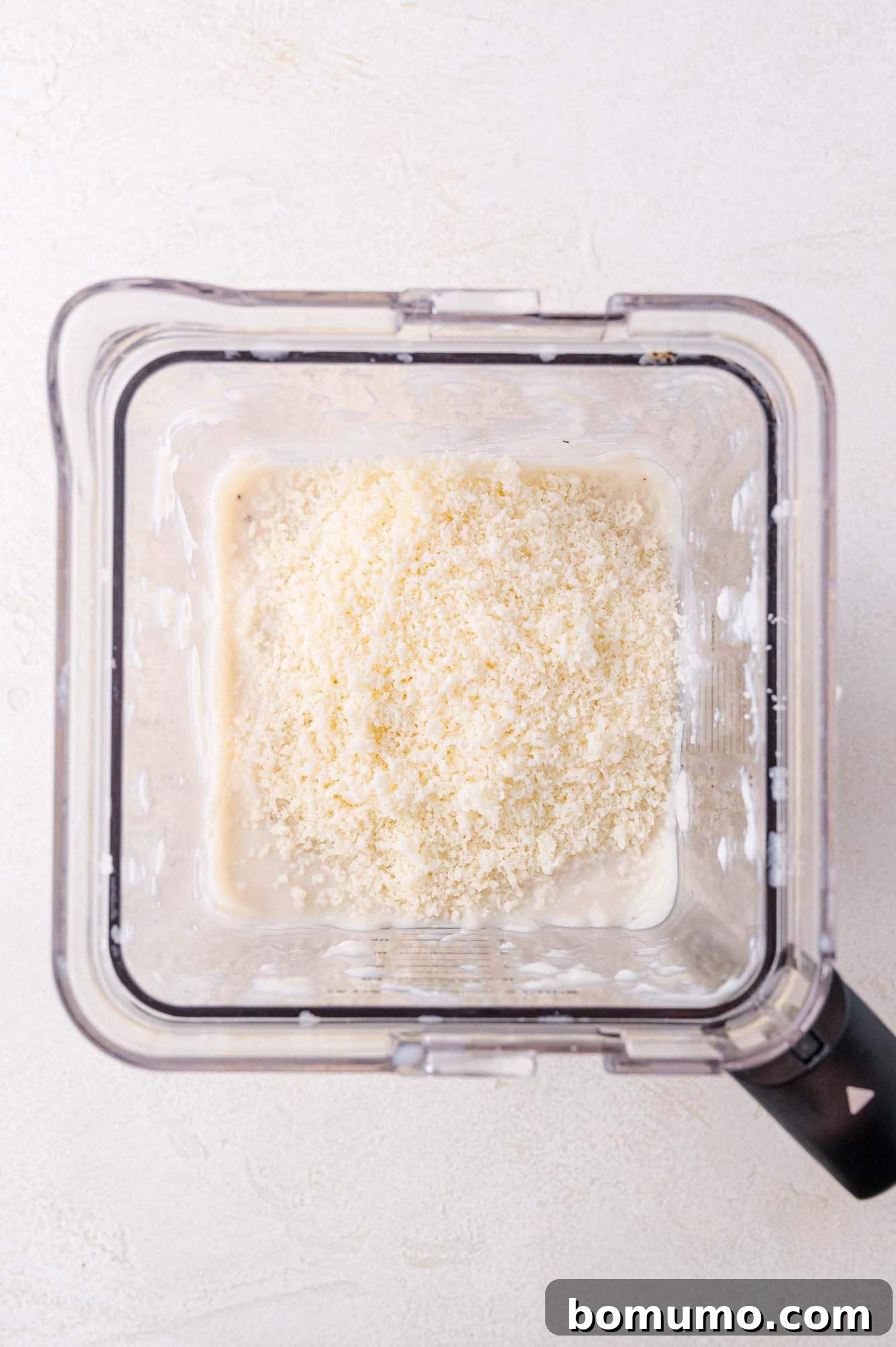 A blender filled with a creamy mixture and finely grated cheese, viewed from above.