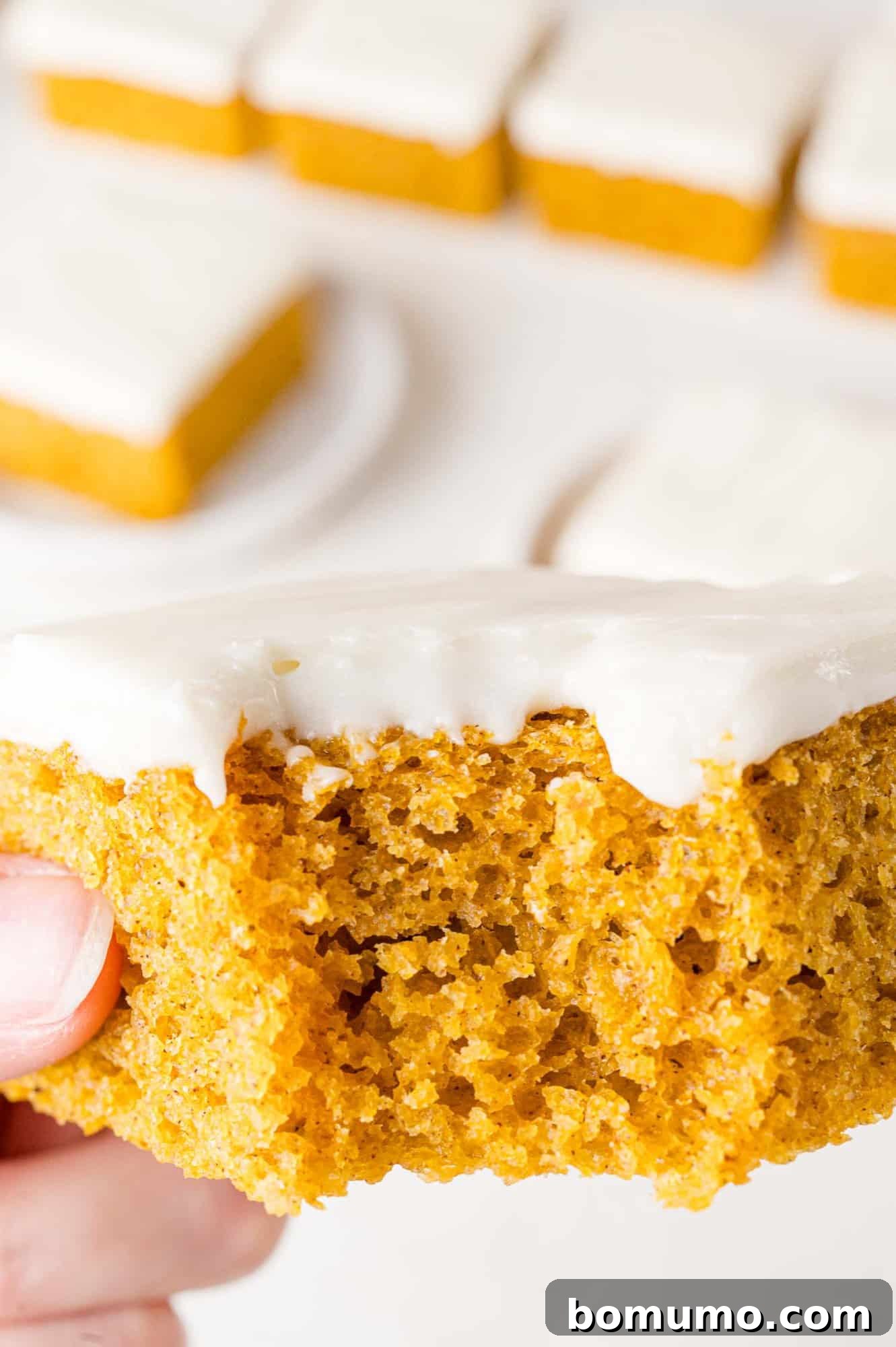 Autumn Pumpkin Spice Bars 18 A hand gently holding a delicious pumpkin bar, freshly frosted and with a tempting bite taken out, revealing its moist interior and inviting texture.
