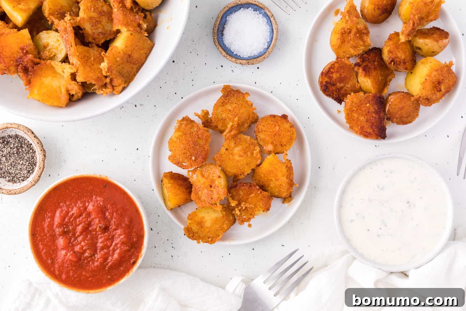 Overhead view of a bowl of crispy parmesan potatoes next to smaller plates, with bowls of ranch and marinara dipping sauces.