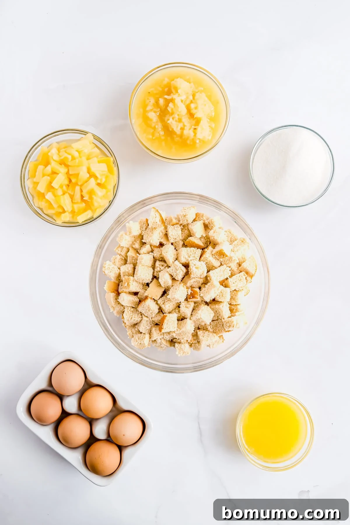 A vibrant overhead shot showcasing all the essential ingredients laid out for making pineapple stuffing: bread cubes, cans of pineapple, sugar, eggs, and melted butter.