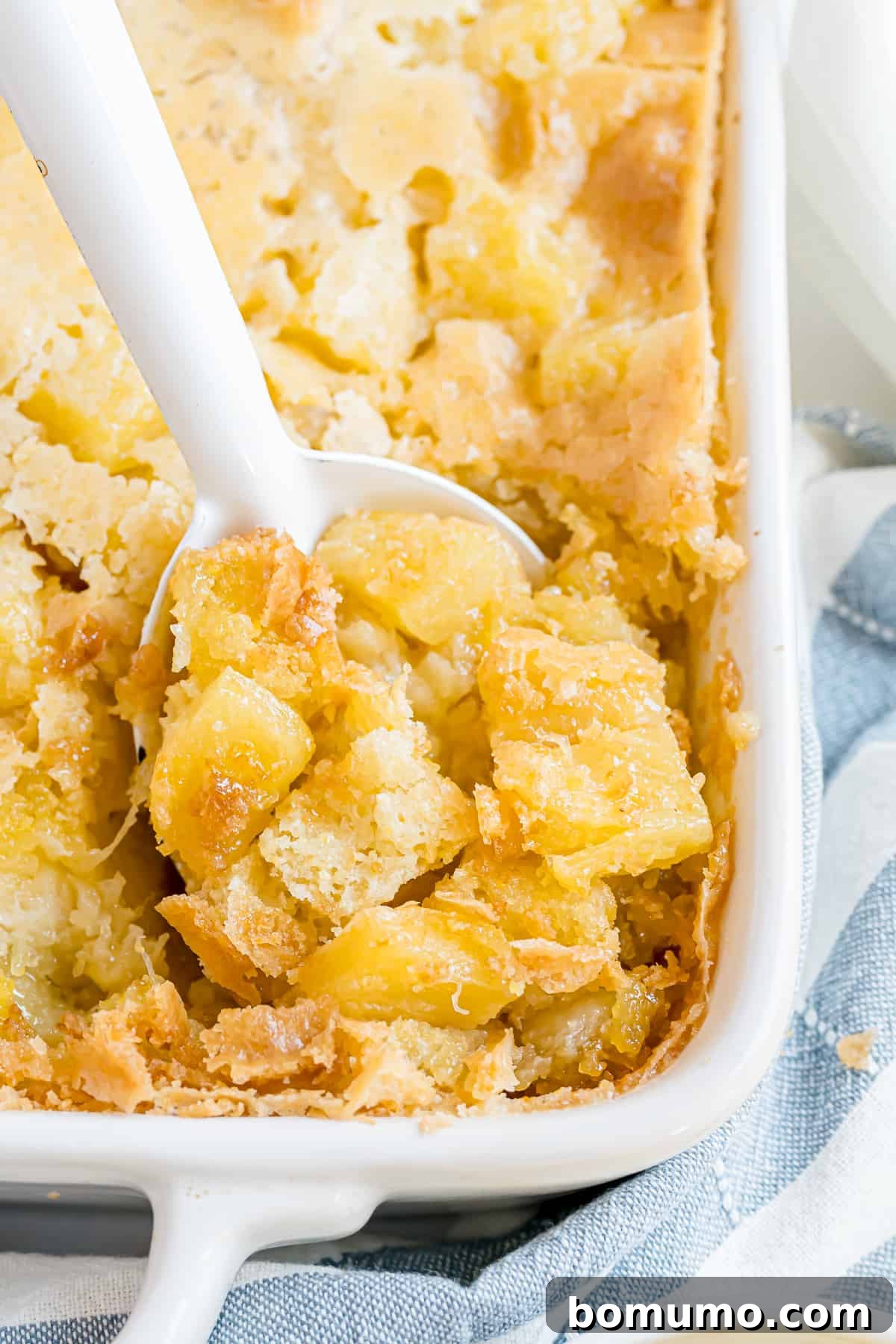 A close-up shot of pineapple stuffing in a casserole dish, with a serving spoon poised to scoop a piece from the golden-brown, bubbly corner.