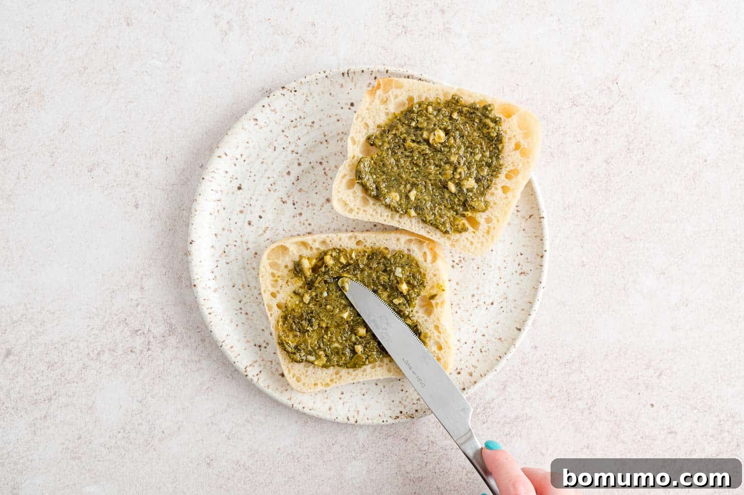 A hand gently spreading green pesto onto a toasted burger bun, adding a layer of fresh flavor.