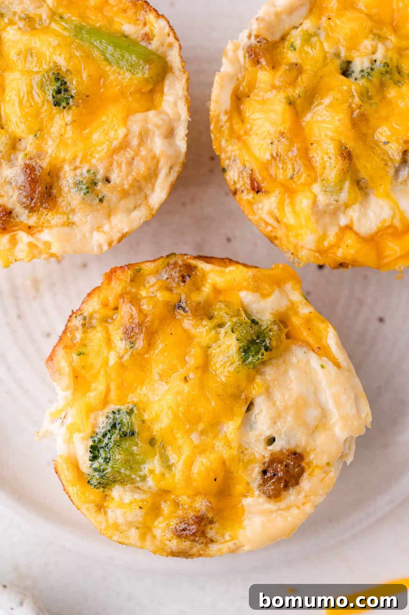 Protein-Packed Egg White Bites 2 Three baked egg white muffins with visible broccoli, sausage, and melted cheddar cheese on a white plate, ready for a healthy breakfast.