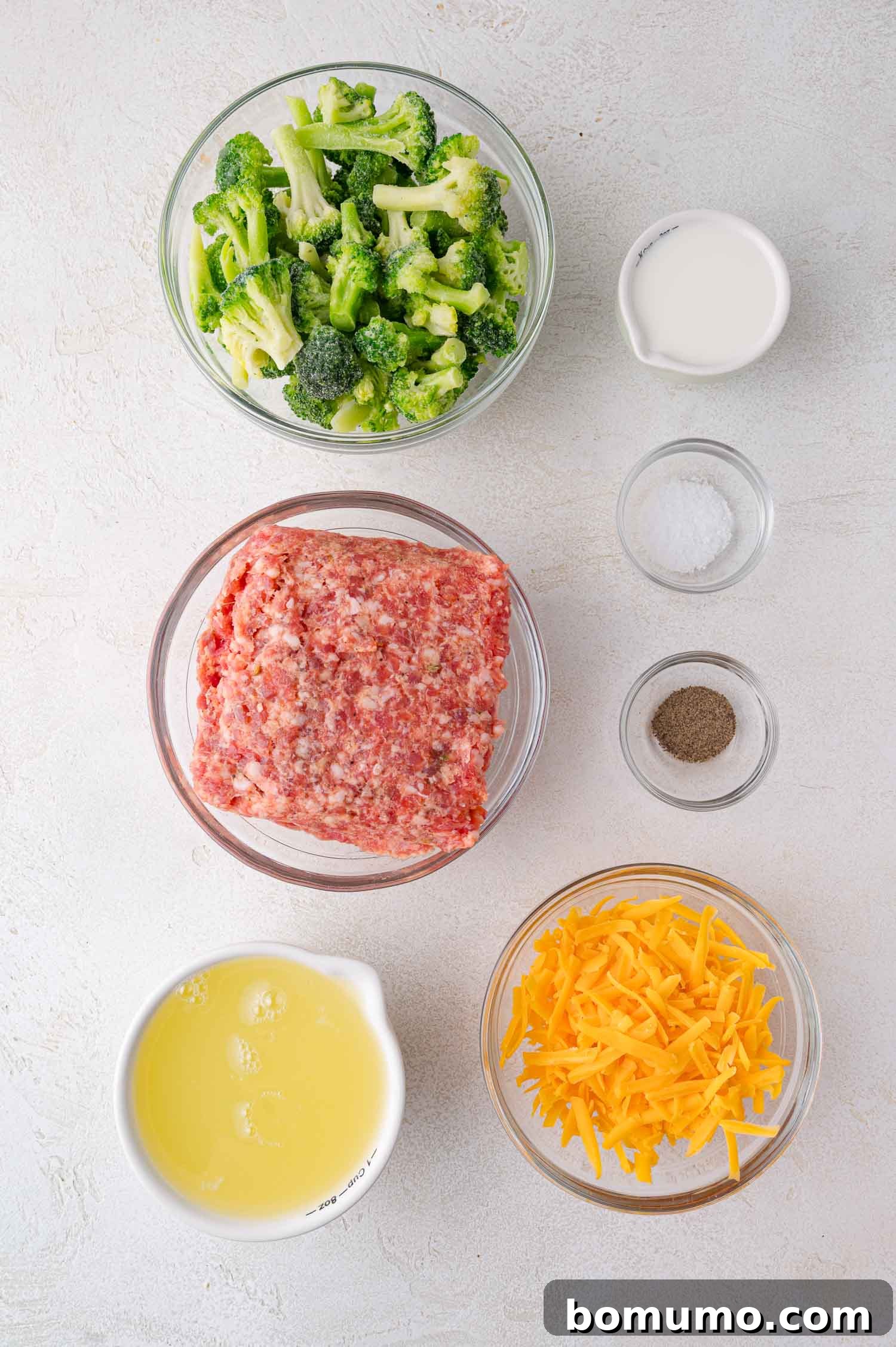 Protein-Packed Egg White Bites 4 Bowls containing chopped broccoli, ground sausage, shredded cheddar cheese, egg whites, milk, salt, and pepper are arranged on a white surface, showcasing all the fresh ingredients needed for the recipe.