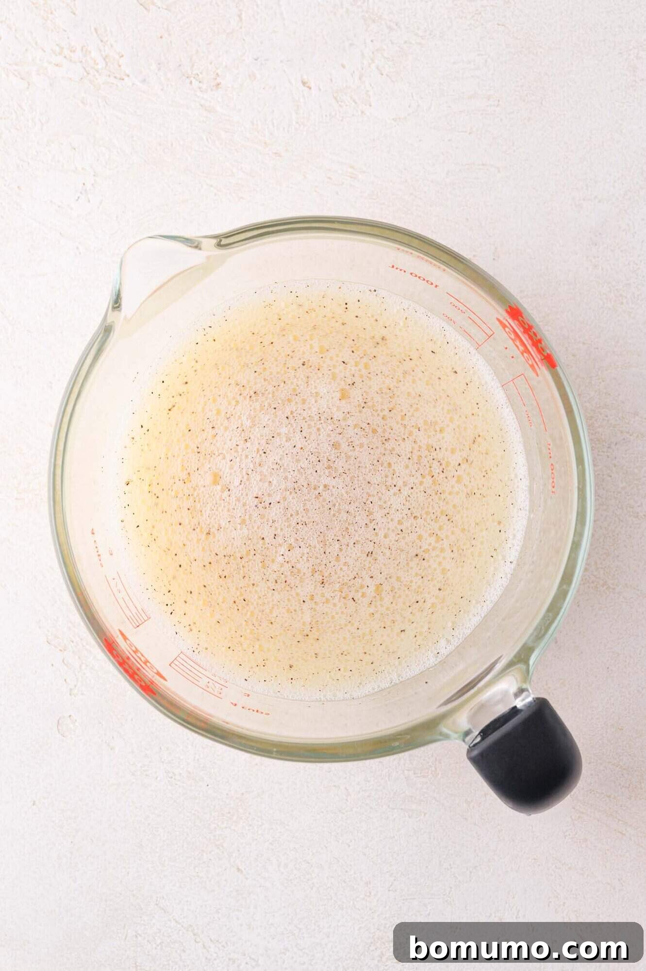 Protein-Packed Egg White Bites 7 A clear glass measuring cup filled with frothy, whisked egg whites on a white textured surface, ready for the next step.