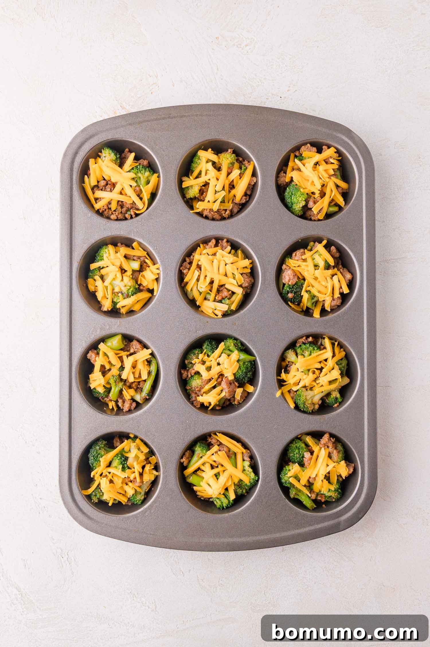 Protein-Packed Egg White Bites 8 A muffin tin with twelve cups, each filled with a base of broccoli, ground sausage, and shredded cheddar cheese, arranged on a light-colored surface, awaiting the egg mixture.