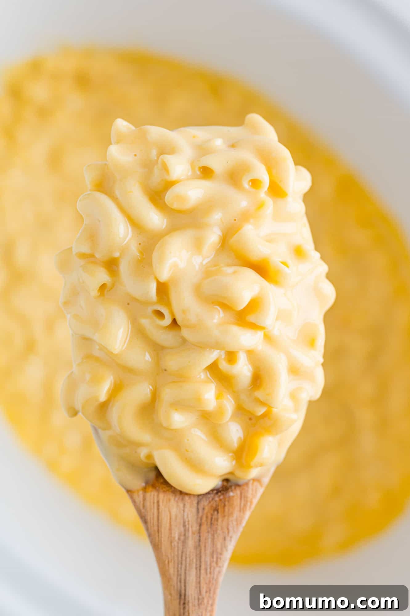 A large serving spoon lifts a generous scoop of golden, rich, and creamy mac and cheese from a slow cooker, showcasing its enticing texture, ready to be served at a party or family meal.