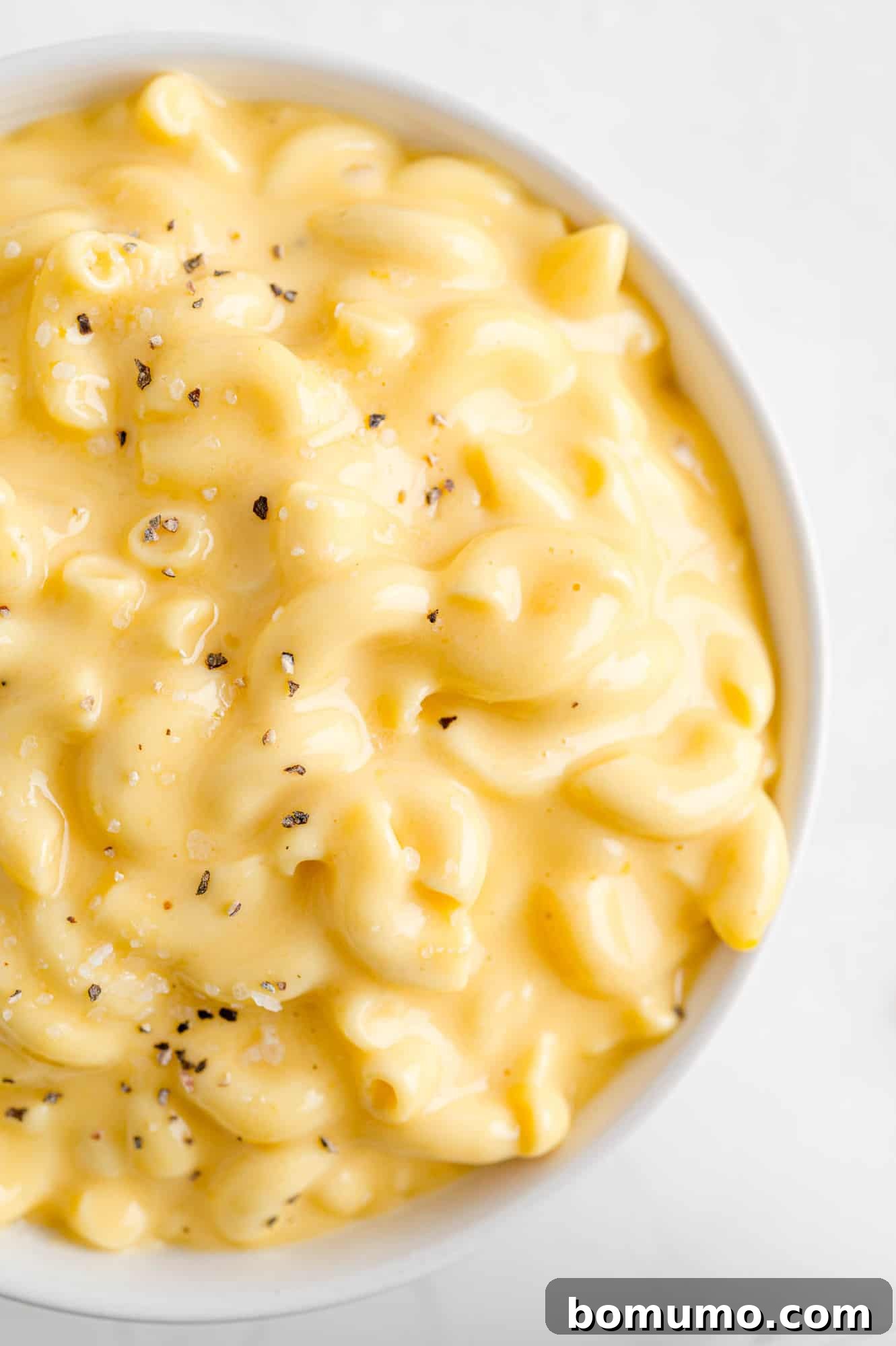 A close-up of creamy macaroni and cheese served in a pristine white bowl, garnished with a sprinkle of fresh chopped herbs, emphasizing its inviting golden hue and smooth, rich texture.