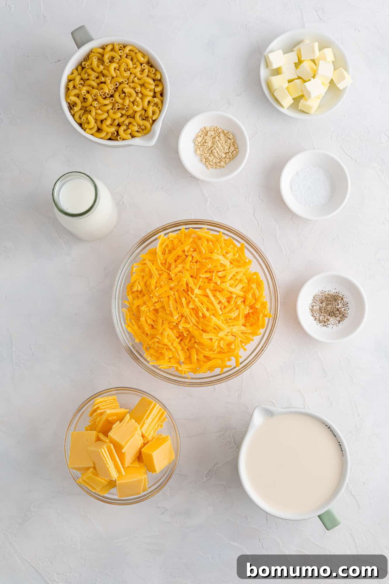 A tabletop display of essential ingredients for making crockpot mac and cheese, including a box of elbow macaroni, bottles of milk, cubed butter, a block of sharp cheddar, and diced American cheese, all ready for preparation.