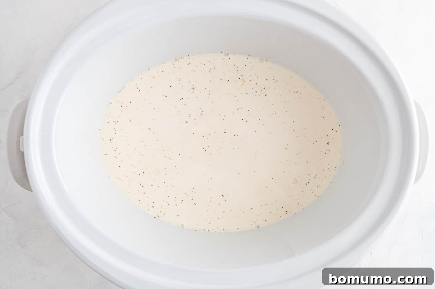 Inside a slow cooker, a homogeneous mixture of whole milk, evaporated milk, and various spices is visible, forming the essential liquid base for the mac and cheese.
