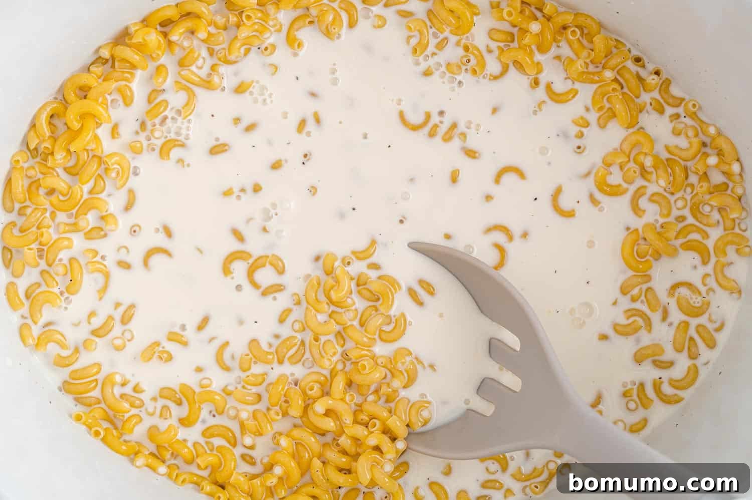 Uncooked elbow macaroni is mixed into a milky liquid base inside a slow cooker, showcasing how the ingredients are combined before the cooking process begins.